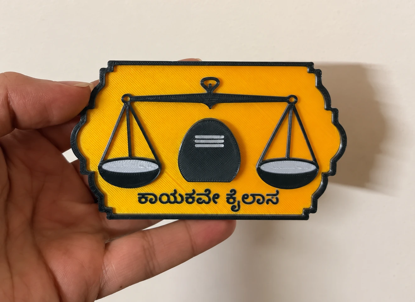 Banajiga Heritage fridge magnet held in a hand, with balanced scales, a linga and the Kannada phrase ಕಾಯಕವೇ ಕೈಲಾಸ